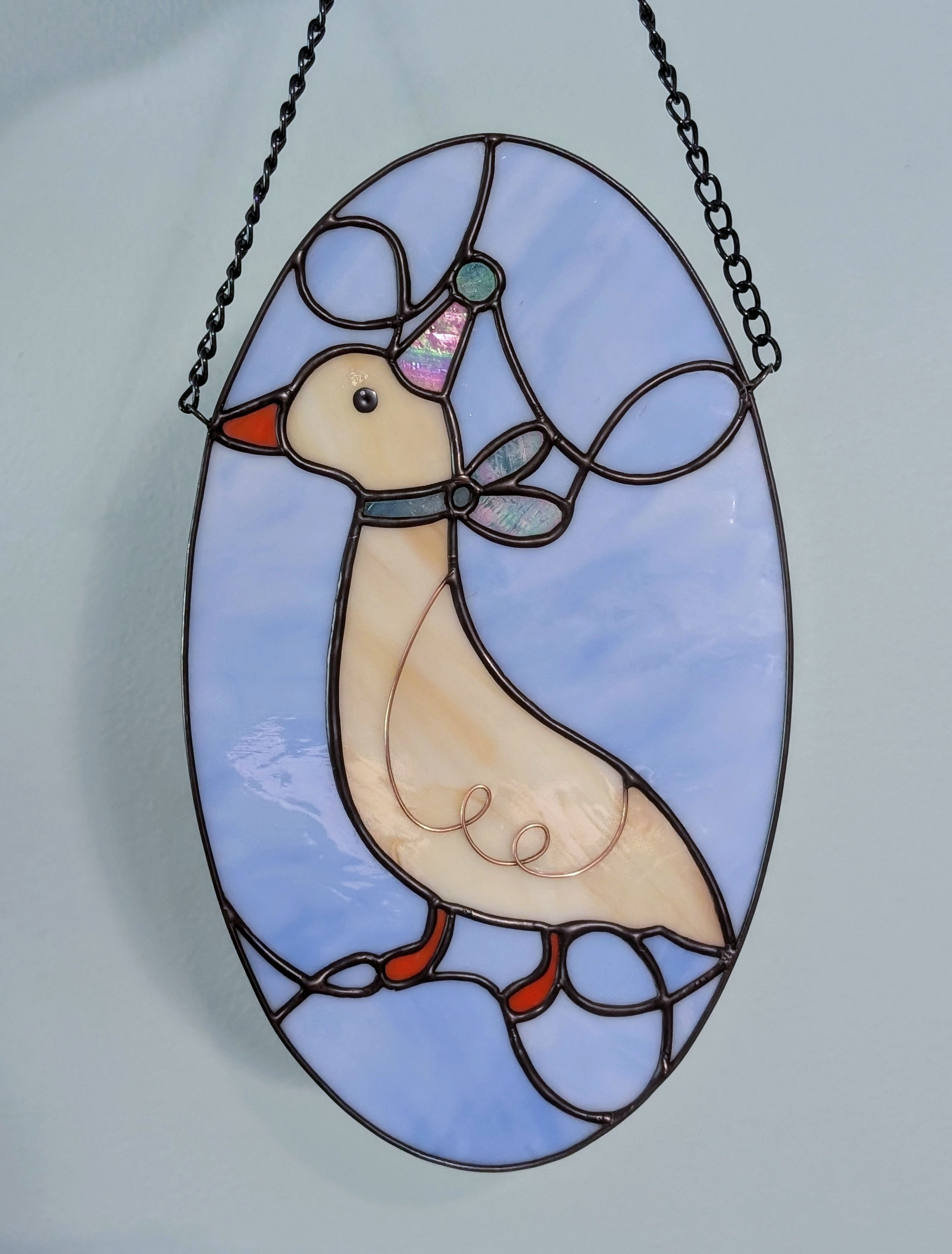 A blue stained glass oval with a white goose with a party hat on
